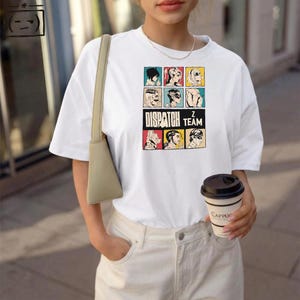 Puede incluir: Camiseta blanca con un gráfico colorido que presenta retratos estilizados y el texto "DISPATCH Z TEAM". La camiseta tiene mangas cortas y cuello redondo. La persona también sostiene una taza de café.