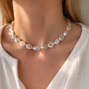 Könnte beinhalten: Eine Perlenkette mit flachen, unregelmäßigen weißen Perlen. Die Kette liegt um den Hals und reflektiert das Licht. Der Hintergrund ist eine weiße Bluse, die eine weiche, elegante Ästhetik erzeugt. Die Perlen sind etwa 1 cm im Durchmesser.
