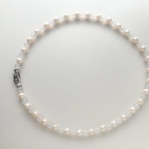 Può includere: Una collana circolare caratterizzata da un motivo alternato di piccole perle sferiche bianche e perline traslucide color bianco sporco. Il filo è fissato con una chiusura a moschettone in metallo argentato, creando un cerchio continuo.
