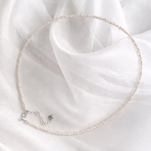 May include: Delicate pearl necklace with a silver clasp and extender chain. The necklace features small, round pearls strung together, creating a simple yet elegant design. The pearls are a creamy white color, and the necklace is displayed on a white fabric background.