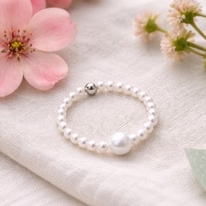 Op de afbeelding: Een delicate parelring met een grote centrale parel en een band van kleinere parels. De ring ligt op een zachte, witte stof, met roze bloemen en groen in de achtergrond.
