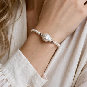 Könnte beinhalten: Ein weißes Perlenarmband mit einem großen, barocken Mittelstück. Das Armband ist mit kleineren, runden Perlen und silberfarbenen Perlen besetzt. Der Schmuck wird an einem Handgelenk vor einem neutralen Hintergrund gezeigt.