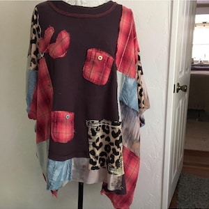 May include: A long-sleeve top featuring a dark brown ribbed knit base with a patchwork design. The garment includes various fabric panels in red plaid, leopard print, and light blue denim, along with two small pockets and buttons.