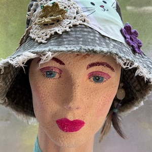 May include: A distressed grey denim bucket hat featuring a frayed brim, a white crocheted lace doily, a small green crocheted flower, a purple crocheted flower, and a light green fabric patch with a printed black and pink cat face design.