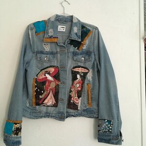 May include: A light blue denim jacket featuring distressed details, a collar, and button-down front. The jacket is decorated with rectangular fabric patches showing figures in traditional clothing with umbrellas, plus blue and floral accent patches.