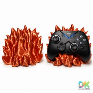 Può includere: Un controller di gioco nero è appoggiato su un supporto a forma di fiamma arancione. È visibile anche un supporto abbinato separato. Il controller ha una luce blu e diversi pulsanti. Il logo "DK MAKES" si trova in basso a destra.