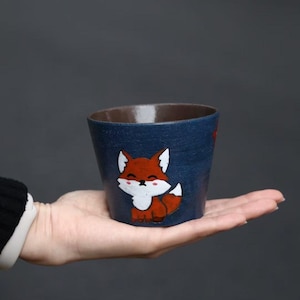 May include: A small, blue ceramic cup with a hand-painted fox illustration. The fox is orange, white, and brown. The interior of the cup is brown. The cup is held in a person's hand against a gray background.