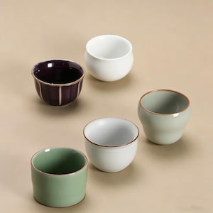 May include: A collection of small ceramic bowls in different colours. The bowls include a deep purple bowl with vertical ridges, a white bowl, a light green bowl, and a white bowl with a gold rim. The bowls are arranged on a neutral background.