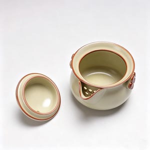 May include: Cream-coloured ceramic teapot with a matching lid. The teapot has a rounded body, a spout with multiple holes, and decorative floral accents. The rim and accents are reddish-brown. The lid is off to the side.