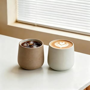 Pode incluir: Duas canecas de café de cerâmica em uma superfície branca. Uma caneca é bege e cheia de café gelado. A outra caneca é branca e contém um café com leite com um desenho em forma de coração na espuma.