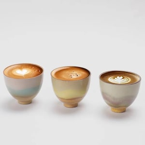 May include: Three ceramic coffee cups with latte art. Each cup has a unique design, with a heart, dots, and a floral pattern. The cups have a rounded shape and a light-colored, speckled finish, with a small base.