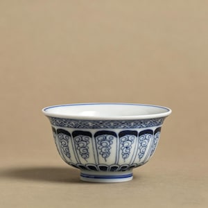 Könnte beinhalten: Eine weiß-blaue Keramikschale mit detailliertem Design. Die Schale hat einen blau gemusterten Rand und ein sich wiederholendes blaues Design auf der Außenseite. Die Schale hat einen kleinen Fuß und steht vor einem neutralen Hintergrund.