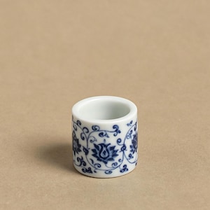 Puede incluir: Un pequeño servilletero cilíndrico de cerámica blanca con un estampado floral azul. El diseño presenta flores estilizadas y espirales. El interior del anillo es blanco liso.