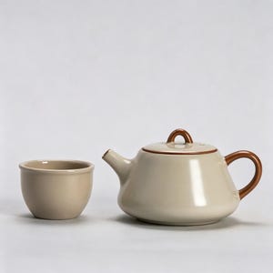 Puede incluir: Una tetera de cerámica color crema con asa, boquilla y tapa marrones. A la izquierda hay una taza a juego. La tetera tiene un cuerpo redondeado y una boquilla pequeña y curvada. La taza tiene una forma simple y cilíndrica.