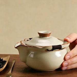 May include: Cream-colored ceramic teapot with a brown rim and lid. The teapot has a unique floral design around the rim and a dark brown handle. A hand is holding the teapot.