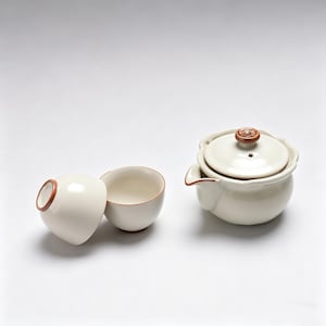 May include: A cream-colored ceramic tea set with a teapot and two teacups. The teapot has a lid with a decorative knob and a spout. The teacups have a rounded shape with a brown rim. The set is designed for brewing and serving tea.