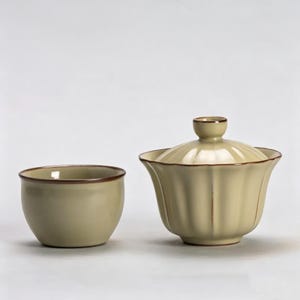 May include: A light green ceramic tea set with a matching cup and a lidded gaiwan. The cup and gaiwan have a brown trim. The gaiwan has a fluted design and a small knob on the lid. The set is designed for tea ceremonies.