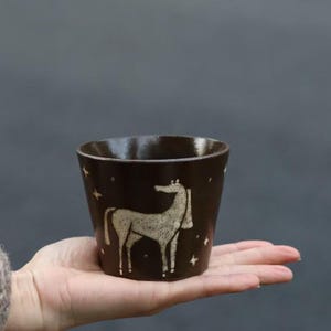 Puede incluir: Una taza de cerámica marrón oscuro con una ilustración de caballo blanco y estrellas. La taza se sostiene en una mano, mostrando su tamaño y diseño. Ideal para bebidas.