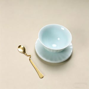 May include: A light blue ceramic teacup and saucer set with a matching gold spoon. The spoon has a unique, curved design. The teacup and saucer have a ribbed design. The set is on a neutral background.