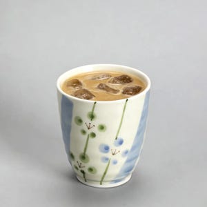Pode incluir: Uma caneca de cerâmica branca com uma bebida castanha clara e cubos de gelo. A caneca tem um design pintado em azul e branco com detalhes florais verdes e azuis. A caneca está em um fundo cinza.