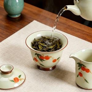 May include: A tea ceremony in progress, featuring a white ceramic cup with a brown rim and a decorative orange fruit design. Hot water is being poured from a white teapot into the cup filled with tea leaves. A matching lid and pitcher are also visible.