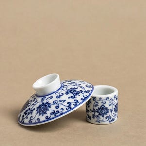Puede incluir: Un juego de taza y tapa de té de cerámica blanca y azul. La tapa tiene un asa y está decorada con un estampado floral azul. La taza es cilíndrica y también presenta el mismo diseño floral azul.