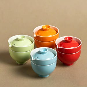 Op de afbeelding: Vier kleurrijke keramische kommen met deksels. De kommen zijn in tinten groen, oranje, rood en blauw. Elke kom heeft een wit interieur en een kleine tuit. De deksels hebben een kleine knop bovenop.