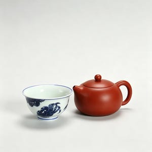 Könnte beinhalten: Eine weiße Keramik-Teetasse mit blauen Wolkenmustern und eine rotbraune Teekanne. Die Teekanne hat eine runde Form mit einem kleinen Griff und einem Deckel mit einem kleinen Knopf. Die Tasse hat einen blauen Rand.