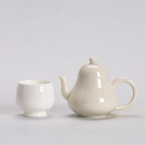 Puede incluir: Una tetera de cerámica color crema con cuerpo redondeado, boquilla curva y asa de bucle, junto a una taza de té de cerámica blanca. La tetera tiene una tapa con un pequeño pomo. Juego de té.