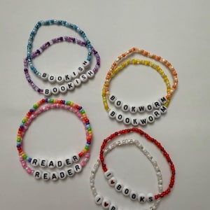 May include: Four beaded bracelets in various colors. Each bracelet features white letter beads spelling out phrases like "BOOKISH", "BOOKWORM", "READER", and "I LOVE BOOKS". The bracelets are a mix of blue, purple, yellow, orange, red, pink, and white beads.
