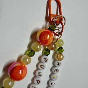 May include: A keychain featuring an orange lobster clasp attached to multiple strands of beads. The strands include white letter beads spelling HELLO PUMPKIN with gold lettering, interspersed with translucent yellow, green, and orange marbled beads.