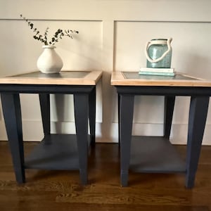 May include: Two matching side tables with navy blue legs and a natural wood top. One table has a white vase with greenery, the other has a glass jar with a rope handle and books. The tables are 61 cm tall.