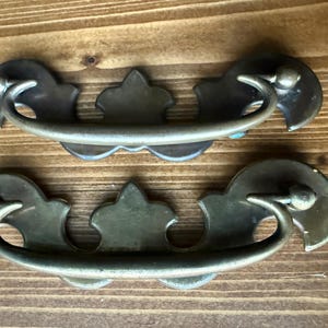 Może przedstawiać: Dwa antyczne mosiężne uchwyty do szuflad z zakrzywionym uchwytem i dekoracyjną płytą tylną. Uchwyty są zamontowane na drewnianej powierzchni z widocznym usłojeniem drewna. Okucia mają estetykę vintage.