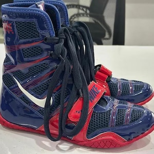 May include: Pair of blue and red boxing shoes with the Nike logo. The shoes have a high-top design with mesh panels, red straps, and white swoosh logos. The soles are red, and the laces are black.