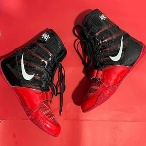 May include: Pair of red and black Nike wrestling shoes. The shoes have a high-top design with a glossy red finish on the lower portion and a black upper with a white Nike swoosh logo. The shoes have laces and a strap closure.
