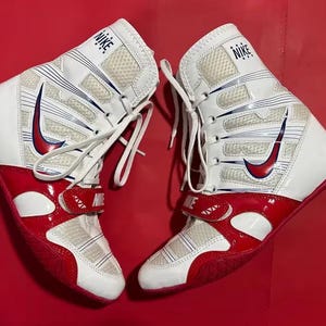 Pode incluir: Par de sapatos de luta Nike brancos e vermelhos. Os sapatos têm um design de cano alto com um logotipo swoosh vermelho e o logotipo Nike na língua. Os sapatos são amarrados e têm uma tira de velcro.