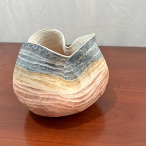 May include: A small, rounded bowl with an irregular, wavy rim. The exterior features horizontal, textured bands of color in shades of slate blue, cream, tan, and terracotta, creating a layered, geological appearance with a matte finish.