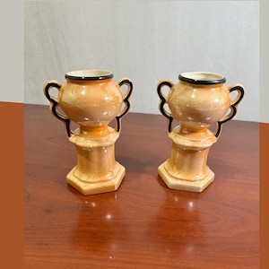 May include: A pair of small, peach-coloured ceramic urns with a glossy finish. Each urn features two curved handles on the sides, a wide opening with a black rim, and a pedestal base. The vessels have a rounded body shape and a smooth, reflective surface.