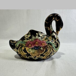 Könnte beinhalten: Eine schwarze Schwanenfigur aus Keramik mit einem handgemalten Blumenkorbmotiv in Rosa-, Grün- und Gelbtönen. Der Schwan ist mit aufwendigen goldfarbenen Verzierungen an Hals, Flügeln und Basis versehen, die die geschwungene Silhouette betonen.