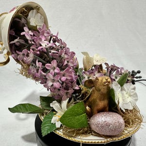 May include: An overturned teacup overflowing with purple hyacinth flowers, white blossoms, and greenery. A golden rabbit figurine and a glittery pink egg sit on the saucer, creating a whimsical Easter-themed display.