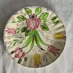 Puede incluir: Plato redondo de cerámica blanquecina con borde festoneado y borde azul punteado. Decorado con un diseño floral central en rosa, verde y amarillo, con flores y hojas estilizadas.