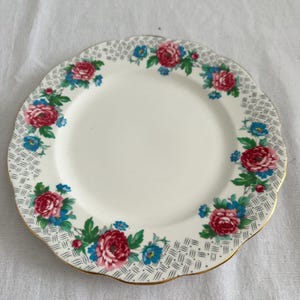 May include: A white ceramic plate with a scalloped edge and a gold rim. The plate is decorated with a floral pattern of red roses, blue flowers, and green leaves. The border has a grey and white woven design.