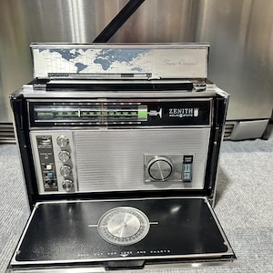 May include: Vintage Zenith Trans-Oceanic radio with a silver and black exterior. The radio features a world map design on the top, a tuning dial, and the Zenith logo. The bottom panel has a compass design. The radio is in a retro style.