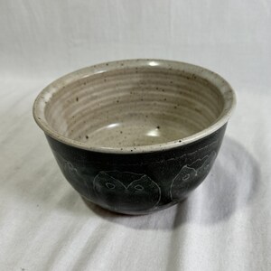May include: A round ceramic bowl featuring a speckled cream-colored interior with visible horizontal ridges. The exterior is finished in a dark charcoal glaze and decorated with a repeating line-art pattern of owl faces encircling the base.