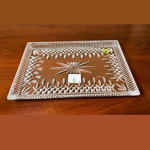May include: A rectangular clear glass serving tray featuring a pressed geometric pattern with a central starburst motif, a cross-hatched border, and a serrated edge. A small green circular sticker and a white rectangular label are attached.