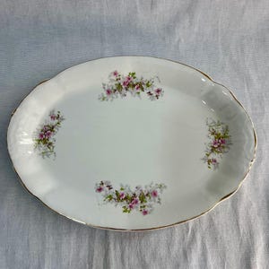 May include: Oval white serving platter with a gold rim and floral design. The platter features clusters of pink and green flowers. The edges have a scalloped design.