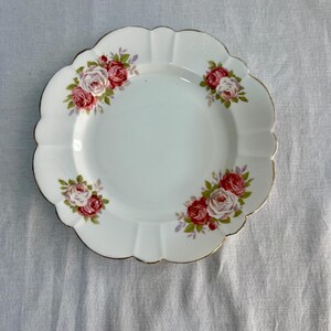May include: White scalloped-edge plate with a gold rim and floral rose pattern. The plate features clusters of red and pink roses with green leaves. A decorative piece for serving or display.