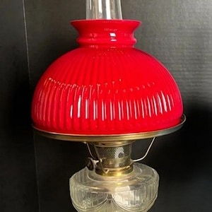 May include: A vintage oil lamp featuring a clear glass base and a bright red glass shade. The shade has a ribbed design and a metal rim. A clear glass chimney extends upwards. This lamp is a classic piece of home decor.