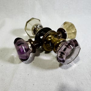 May include: Two vintage door knobs featuring faceted glass handles, one clear and one purple, connected by a central brass mechanism with circular mounting plates. The hardware includes dark metal collars securing the glass to the brass spindle.