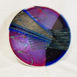 May include: A round ceramic plate featuring a vibrant gradient of magenta, purple, and deep blue. The surface is decorated with a textured, metallic-toned diagonal band and intricate, thin, dark lines that create a layered, abstract pattern.